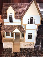 Front view of the wooden dollhouse showing shingle roof, porch, staircase, and window details.