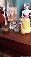 Wedgwood figurine of a girl in colorful dress, glass fish trinket box, and small teddy bear quartz clock on wooden table.