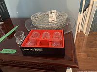 Six Cristal Darques crystal liquor glasses in original box on wood surface, three glass platters stacked behind