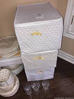 White quilted storage box with three drawers. Three crystal glasses placed in front of the box on floor.