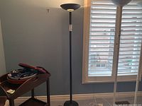 Black pole lamp standing near a window with white blinds, showing the full height and design of the lamp and shade.