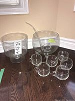 Glass punch bowl, ladle, and six cups on dark wood floor near corner.