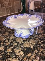Front side angled view of the Shelley Dainty Blue cake plate on granite countertop, showcasing the delicate blue floral pattern and scalloped edges on both the plate and pedestal base.