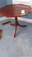 Full view of wood occasional table with scalloped round top, tripod legs, and casters on a carpeted floor.