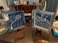 Two square ceramic plates on stands displayed on a table, showing overall size and artwork.