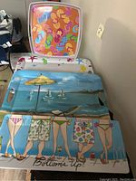 Photo shows four colorful beach-themed plastic serving trays stacked together. The bottom tray depicts legs in swimwear and the ocean background with beach chairs and a yellow umbrella. The top tray has a colorful flip-flop pattern on white with an orange base.