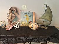 Wide shot showing all five beach themed decorative items: driftwood seahorse, welcome slate sign, ceramic fish, metal sailboat, and jar with sand and shells.