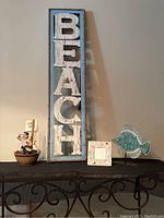 Four decorative beach-themed items displayed together: sailor figurine in basket with mast, vertical BEACH sign, 6x6 picture frame with ceramic fish, and turquoise metal and ceramic fish figurine on stand.