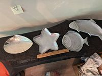Four pewter metal serving dishes displayed together on dark textured surface; clam shell, star, and two fish shapes visible