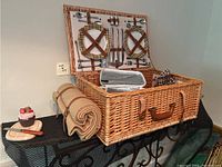 Open wicker picnic basket showing plaid lined interior with leather strapped plates, cutlery, salt and pepper, and wine opener attached to lid.