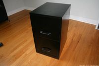 Black 2 drawer metal filing cabinet standing on a hardwood floor, showing exterior from an angle.