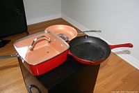 Cast iron skillet with red handle alongside square and round copper-finish pans with stainless steel handles and matching lid on square pan.