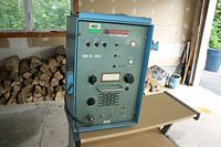 Front and side view of the Spilsbury & Tindal Model MRT-700 radio transceiver unit placed on a cart in a garage showing wood logs in background.