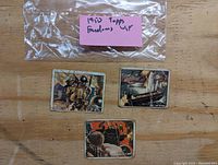 Photo showing three 1950 Topps Freedom's War trading cards with a label in background. Cards feature wartime illustrations; two with grey backs and one tan back.