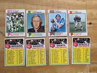 Photo showing four football player cards (Merlin Olsen, Fred Biletnikoff, Greg Landry, Mike Stratton) and four corresponding team checklist cards for Raiders, Browns, Saints, Bengals arranged on a wooden surface.