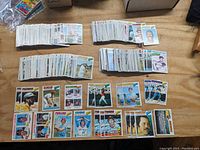 Photo showing four stacks of baseball cards arranged on a wooden surface, along with a spread of a few cards face up featuring various players and card designs from the 1977 Topps set.