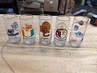 Front view of all five vintage NCAA drinking tumblers lined up on wooden surface showing their logos and mascots clearly.