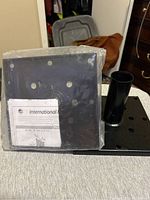 Photo of two black metal fence post brackets, one in plastic packaging with instruction sheet, the other unpacked showing base plate and cylindrical post holder.