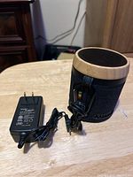 Photo shows cylindrical House of Marley speaker with black fabric and wood top ring, power adapter and audio cable on table.