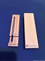 Meater+ wireless Bluetooth BBQ thermometer with wooden charging dock and box on blue background.