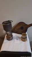 Antique fireplace smoker blower, large ribbed metal topped miners lamp, and smaller brass-style miners lamp displayed on white cloth.