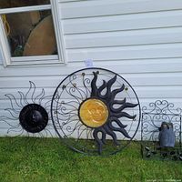 Photo showing the full set of three black metal garden wall decorations including two sun faces and one owl panel on grass by white siding