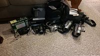 Multiple cameras, video cameras, small tripods, carrying cases, and film box arranged on carpet.