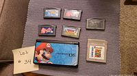 Photo showing Nintendo DS console with 5 game cartridges laid out on a carpet surface, some damage visible on the labels of cartridges.