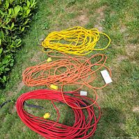 Three heavy-duty extension cords arranged on grass featuring yellow, orange, and red colors with plugs visible.