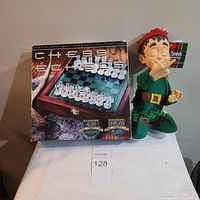 Lot showing battery operated Grumpy Elf figurine and original box of glass chess set on white surface.