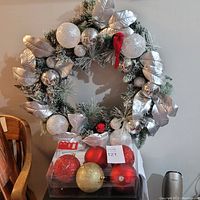 Image showing the entire wreath decorated with silver leaves, white and silver balls, and a red bird on a wall beside packaged large Christmas balls in red, gold, and textured finishes.
