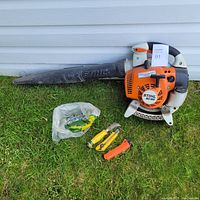 Full view of the STIHL BG86 leaf blower lying on grass next to the pruning knife, hand saw, utility knife, and bag of hardware.