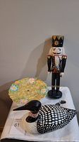 Photo showing ceramic loon, nutcracker figurine in background, and floral glass plate.
