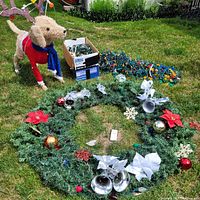 Photo of the full Christmas wreath decorated with ornaments and ribbons, the box containing light strings, and the assembled lighted dog figure with scarf and antlers on grass.