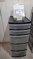 Full image of the tall plastic drawer unit with translucent drawers and black top, with basket on top containing plastic furniture covers.