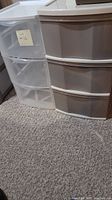 Two plastic drawer units side by side on carpeted floor, one clear and one grey.