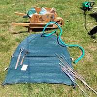 Wooden garden wagon visible in background, green shrub covers laid on grass with metal wire frames, blue garden hose coiled on top, hand cultivator and rake laying on shrub covers, bundle of metal stakes beside shrub covers.