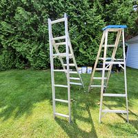 Photo showing two aluminum ladders standing on grass: one extension ladder and one A-frame step ladder with blue top.