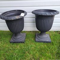 Two large black garden pots shown side by side outdoors on grass, against a white siding wall. Both pots have a fluted urn shape and square bases.