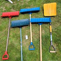 Five winter shovels with plastic blades in different colors lying on grass, showing the overall variety and condition.