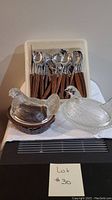 Full lot with vintage cutlery set in white holder behind two nesting hen glass covered dishes (one clear brown ceramic base, one fully clear glass) placed on white cloth