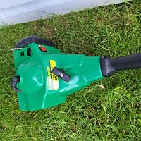 Top view close-up of green gas-powered weed wacker showing X-T label, on/off switch, and black handle on grass