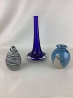 Photo showing three vases including tall blue glass vase, small blue and cream pottery vase, and gray and white pottery vase.