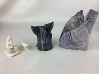 Three collectible hand made carvings displayed on a white surface: a small light colored figure carving with an animal, a dark abstract animal head carving, and a tall abstract stone carving with green and purple hues and incised lines.