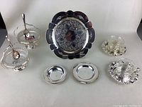 Front view of all silverplated servingware items arranged including a large tray, two small dishes, two glass bowls with spoons, and two glass cup and saucer sets.