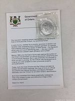 Front view of the coin in original plastic packaging attached to card showing Government of Ontario crest and description about the coin's symbolism.
