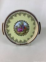 Full view of round Bavarian china plate with handles and detailed painted scene in center, floral border and gold trim.