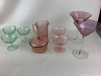 Full display of all vintage colored glassware pieces on white surface including green sherbet glasses, pink pitcher, sugar bowl, cordial glasses, and purple martini glasses