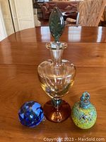 Clear blown glass amphora with stopper, blue glass ball, and small colorful glass piece on wood table.