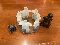 View of seven frosted glass Inuit-style figures arranged in a circle on a round glass plate alongside a dark stone figurine; one glass figure is broken and needs regluing.